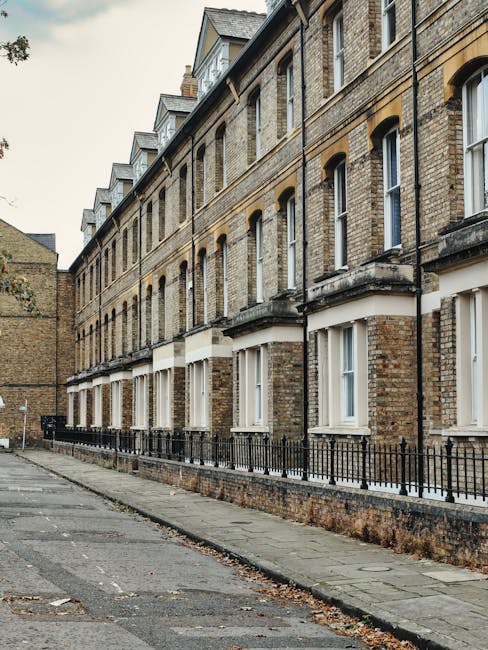 The image depicts the exterior of a row of traditional brick residential buildings in Brixton, SW2, featuring multiple levels with sash windows and small balconies. The brick façade appears aged but well-maintained, with a beige and brown color palette. The sidewalk in front is paved with concrete slabs, showing some fallen leaves and minor debris. A black metal railing runs along the front of the buildings, separating the pathway from the ground-floor windows. The lighting suggests an overcast day, creating a neutral tone across the scene. For property owners seeking professional deep cleaning and surface sanitisation, Cleaners SW2 offers expert services to enhance and maintain the cleanliness of similar residential exteriors, ensuring a hygienic environment and preserving building appearance.