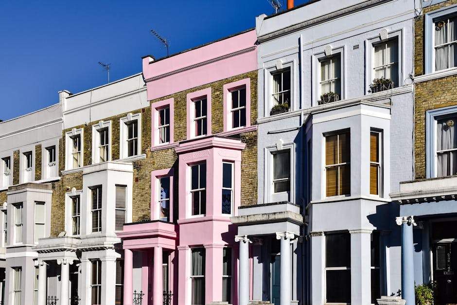 A row of Victorian-style terraced houses in Brixton, SW2, featuring a mix of white, pastel pink, and light yellow facades with large sash windows, some with decorative moldings and window planters. The buildings are set against a clear blue sky, with sunlight casting shadows on the exteriors. The image highlights the architectural details and varying textures of brick and painted surfaces, illustrating the charming, historic urban residential environment. Cleanliness and well-maintained appearances are implied, aligning with the context of professional domestic and end of tenancy cleaning services offered by Cleaners SW2.