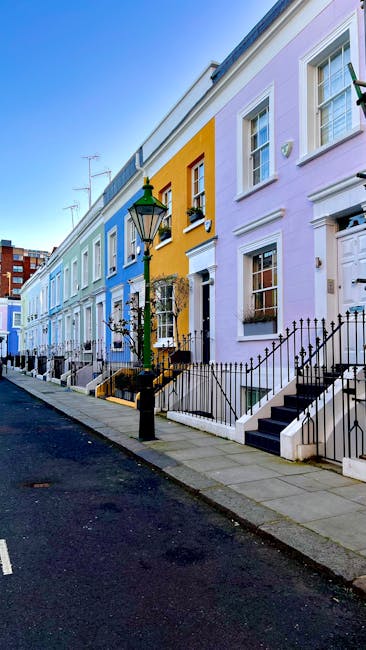 The image depicts the exterior of a row of traditional brick residential buildings in Brixton, SW2, featuring multiple levels with sash windows and small balconies. The brick façade appears aged but well-maintained, with a beige and brown color palette. The sidewalk in front is paved with concrete slabs, showing some fallen leaves and minor debris. A black metal railing runs along the front of the buildings, separating the pathway from the ground-floor windows. The lighting suggests an overcast day, creating a neutral tone across the scene. For property owners seeking professional deep cleaning and surface sanitisation, Cleaners SW2 offers expert services to enhance and maintain the cleanliness of similar residential exteriors, ensuring a hygienic environment and preserving building appearance.