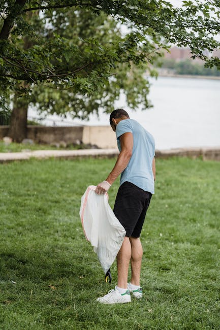 A person is engaged in a park cleanup activity, wearing a light blue t-shirt, black shorts, and white sneakers. They are holding a large white trash bag and appear to be collecting litter from the grassy area near a tree with dense green foliage. The park setting includes well-maintained green grass, a paved pathway, and a water body in the background under natural daylight. This scene highlights community effort in maintaining cleanliness in an outdoor space, consistent with professional cleaning and sanitation efforts promoted by Cleaners SW2, especially in relation to outdoor environmental hygiene.