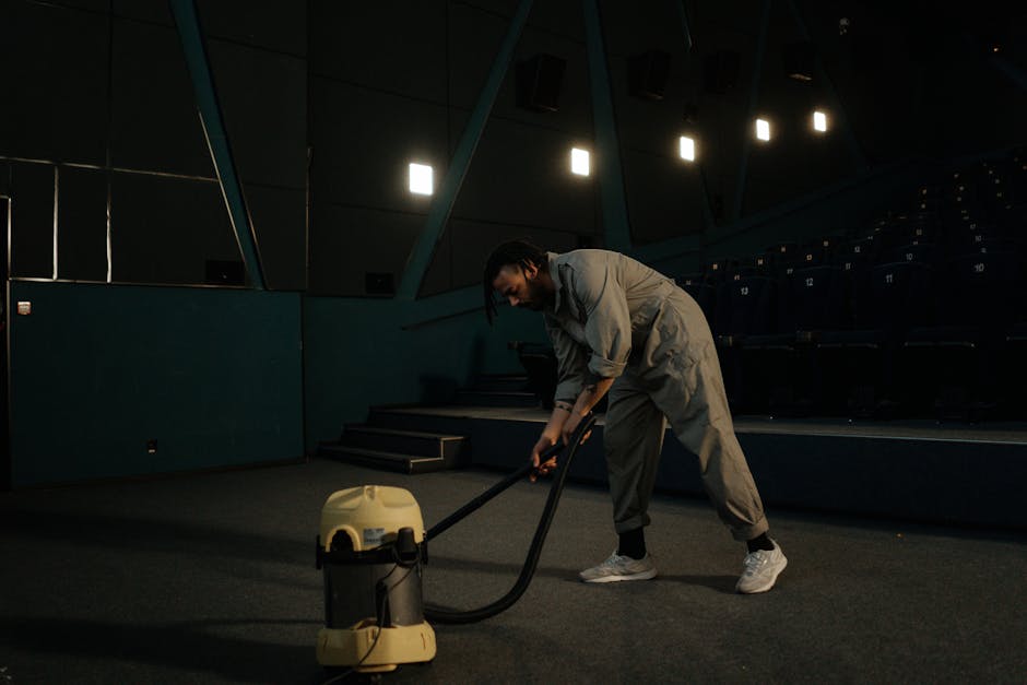 A person operating a commercial vacuum cleaner with a yellow and black body, vacuuming a dark carpeted floor in an empty auditorium or theatre with navy blue seating and a dark, acoustically treated wall. The individual is dressed in light-colored overalls and sneakers, leaning forward slightly while focusing on thorough surface cleaning. Overhead, bright square and rectangular lights illuminate the space, reflecting off the smooth, dark surfaces and emphasizing the clean, dust-free condition of the environment. The setting suggests a professional approach to deep cleaning and maintenance, consistent with services offered by Cleaners SW2 for ensuring hygienic and well-maintained commercial spaces, as highlighted on the Coldharbour Lane carpet cleaning specialists Brixton, SW2 page.