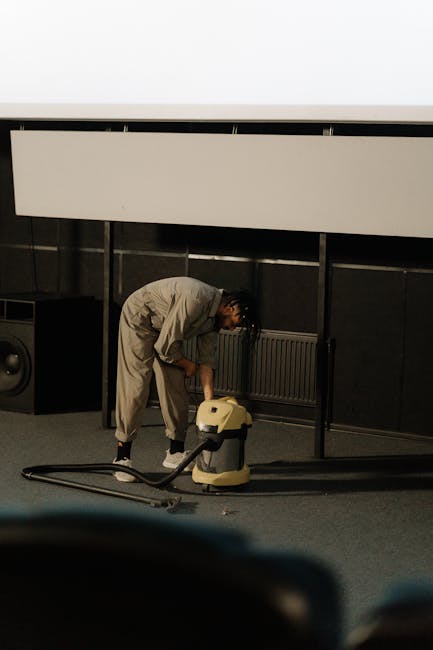 A person wearing light-colored overalls is operating a vacuum cleaner on a carpeted floor in a room with dark walls and a large white projection screen mounted on the wall. The individual appears to be engaged in deep cleaning or surface cleaning as part of a professional cleaning service, with equipment placed nearby. The room has a modern appearance with minimal decor, and the lighting suggests an indoor, controlled environment. This scene exemplifies thorough carpet cleaning, a key aspect of domestic cleaning services offered by Cleaners SW2 on Coldharbour Lane, Brixton, SW2.