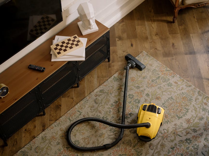 A person operating a commercial vacuum cleaner with a yellow and black body, vacuuming a dark carpeted floor in an empty auditorium or theatre with navy blue seating and a dark, acoustically treated wall. The individual is dressed in light-colored overalls and sneakers, leaning forward slightly while focusing on thorough surface cleaning. Overhead, bright square and rectangular lights illuminate the space, reflecting off the smooth, dark surfaces and emphasizing the clean, dust-free condition of the environment. The setting suggests a professional approach to deep cleaning and maintenance, consistent with services offered by Cleaners SW2 for ensuring hygienic and well-maintained commercial spaces, as highlighted on the Coldharbour Lane carpet cleaning specialists Brixton, SW2 page.